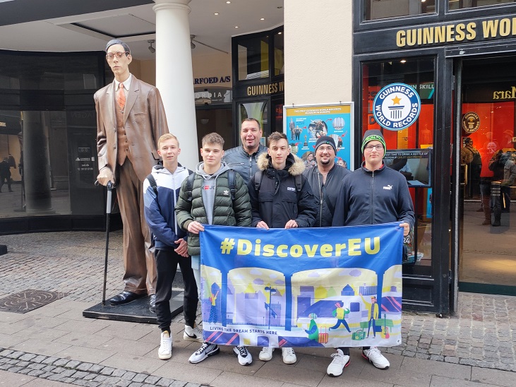 Fiatalok egy csoportja egy sétálóutcában DiscoverEU feliratú zászlót tart a magasba