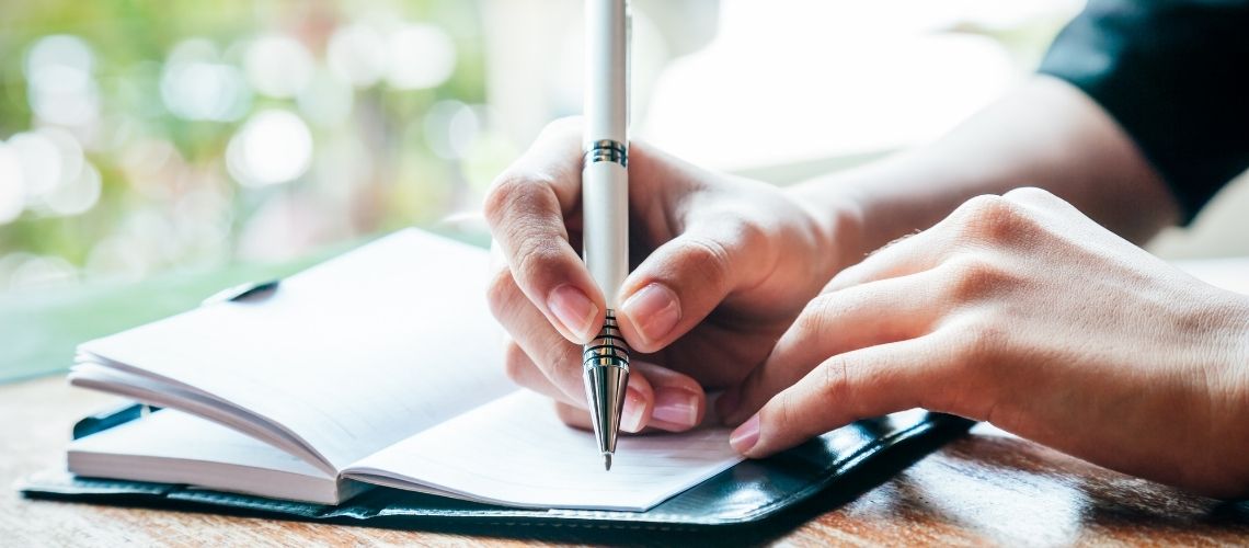 Határidőnaplóba író kéz egy napsütötte asztalon