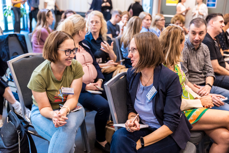 Két hölgy egy konferenciateremben székeken ül és egymással beszélget