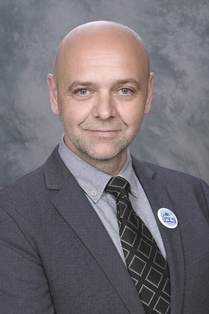 Öltönybe és nyakkendőbe öltözött férfi szürke háttérrel kitűzővel a ruháján mosolyog