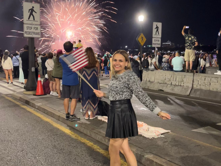 Fiatal egyetemista lány egy úttest közepén áll kezében USA zászló háttérben tűzijáték