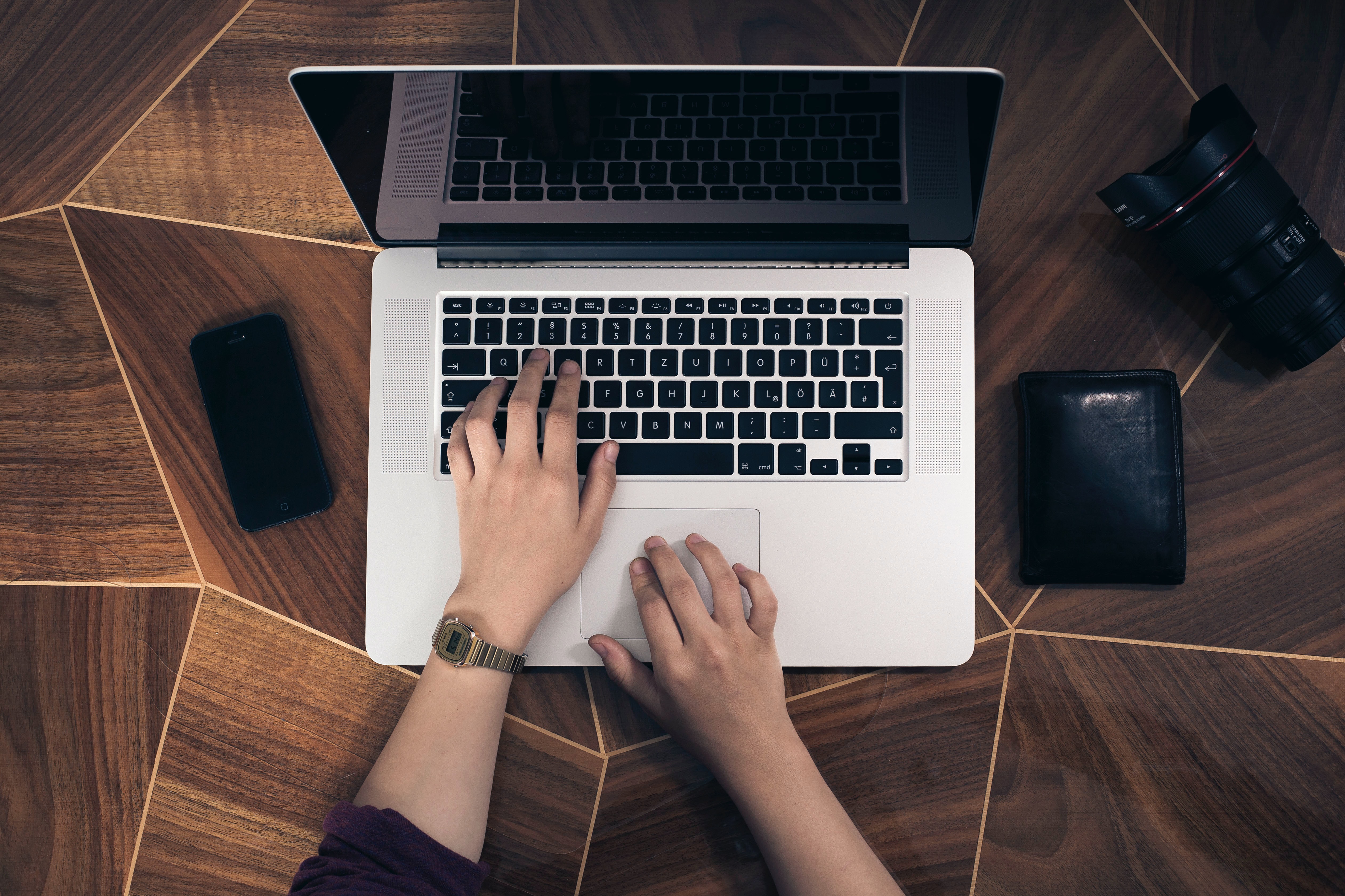 Egy faberakásos asztalon látható egy laptopon dolgozó ember keze és tárcája telefonja és a fényképező teleobjektívje