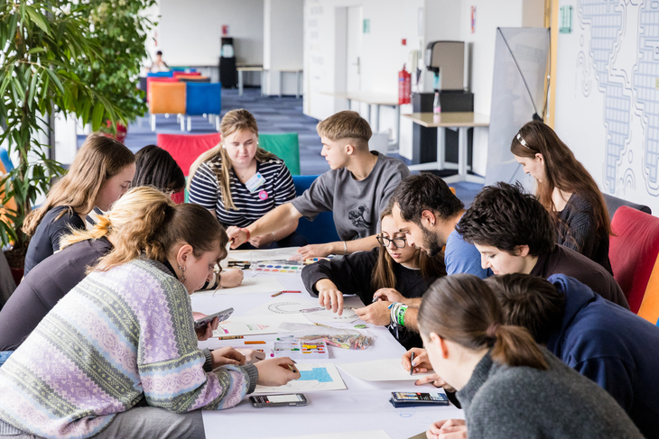 DiscoverEU After esemény résztvevői egy asztal fölé hajolva ülnek körben és alkotnak 