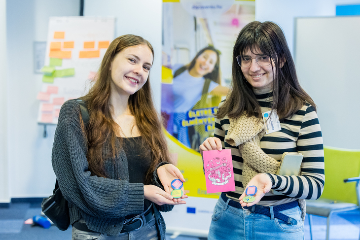 2 fiatal DiscoverEU After résztvevő tartja a kezében és mutatja a kamerának a képeslapokat és kitűzőket