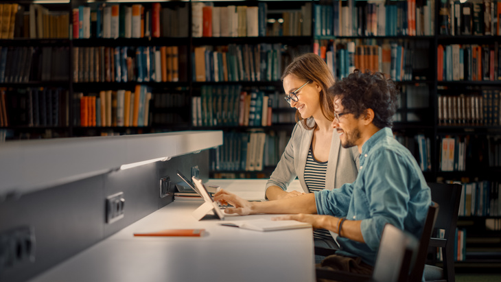 Két felnőtt olvas egy asztalnál a könyvtárban