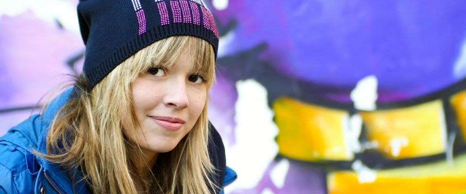 Szőke fiatal kék kabátban és sapkában félprofilból néz szembe a kamerával