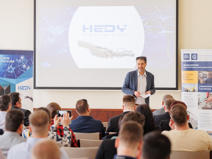 Kivetítő előtt öltönyös előadó a HEDY egyetemi projektről tart előadást