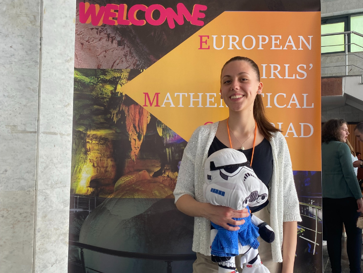 Pannónia Ösztöndíjas diáklány áll egy női matematika versenyt hirdető tábla és egy épület előtt