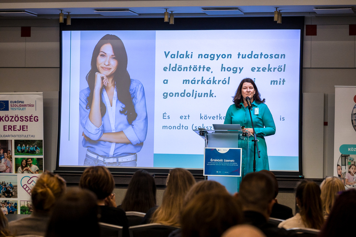 Barna hajú hölgy előadást tart mögötte kivetítőn egy női kép és egy felirat