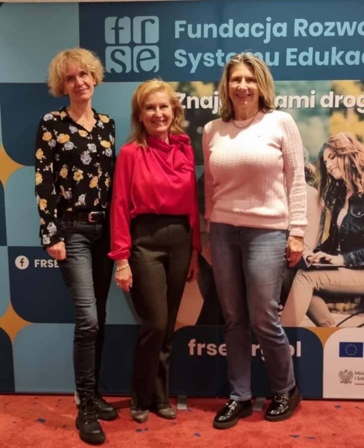 Három hölgy fényképezkedik egy oktatási konferencia plakátja előtt