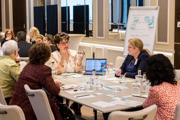 Világos teremben egy asztal körül négy ember ül és az előadóra figyel