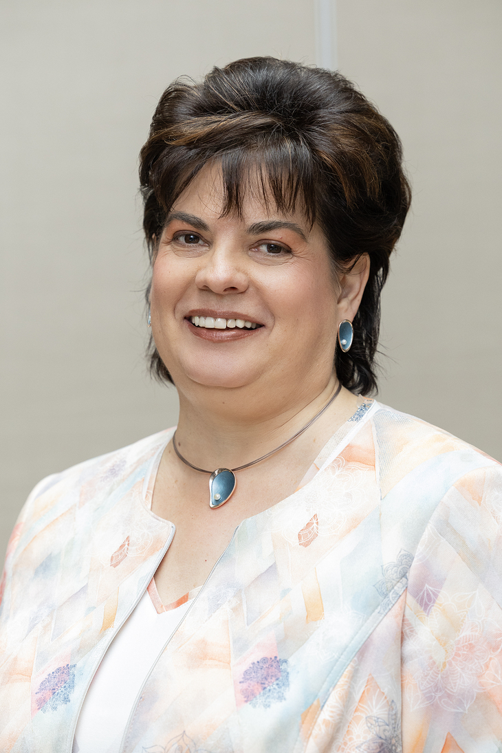 Barna hajú hölgy profilfotója aki világos zakóban kék nyakláncot és fülbevalót visel