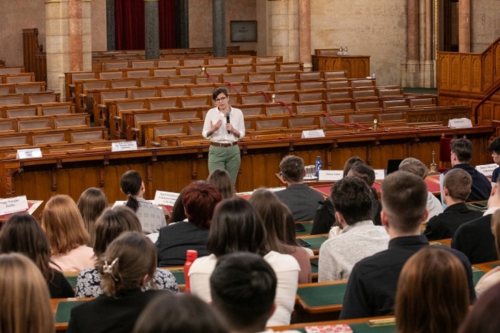 Orosz Anna országgyűlési képviselő mikrofonnal a kezében beszédet tart a parlamenti székeken ülő diákok előtt