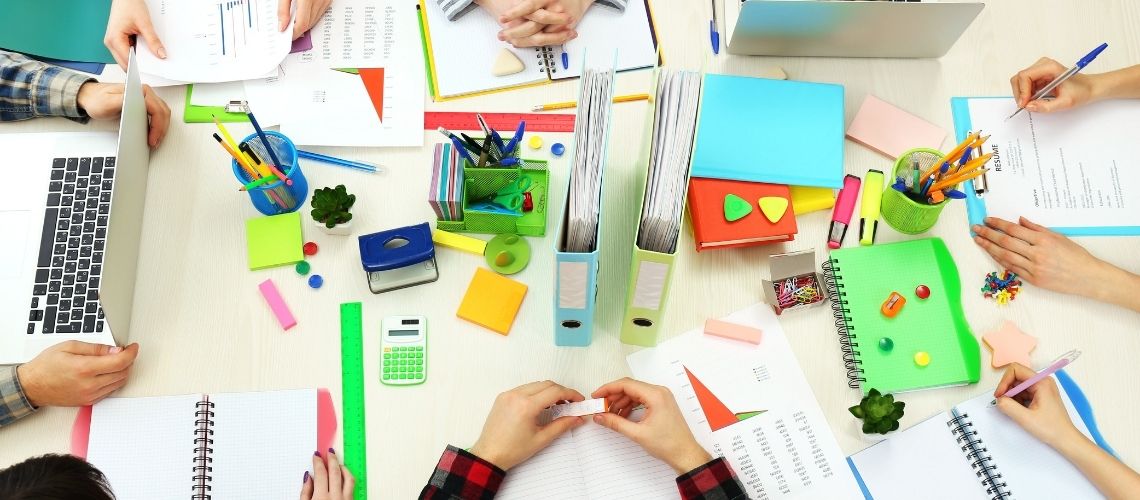 Fiatalok kezei látszanak egy asztal körül amelyen különféle színes irodaszerek hevernek