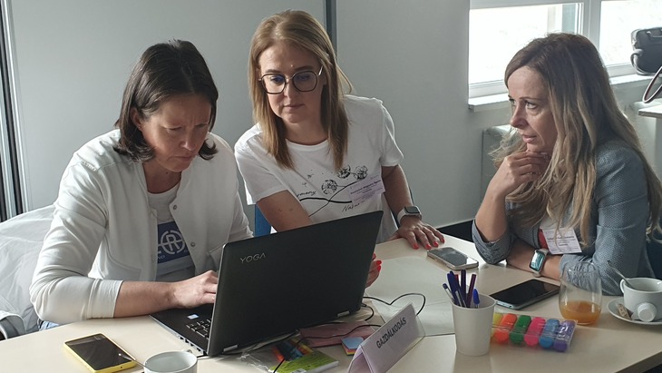 Projektoktatás workshop 3 résztvevője egyeztet egy íróasztalon a laptop körül
