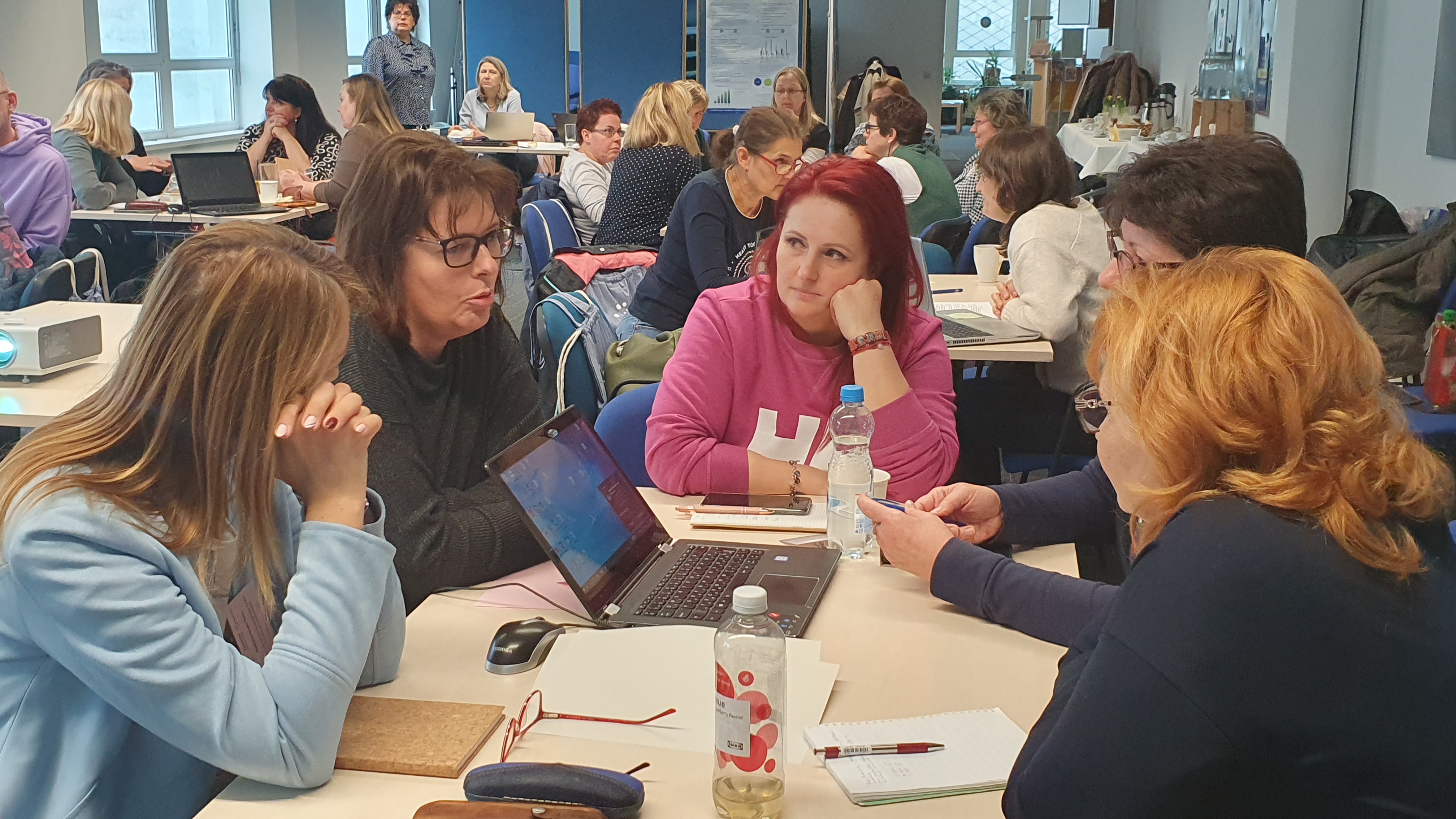 akkreditált oktatók csapatai ülnek egy konferenciateremben több asztalnál
