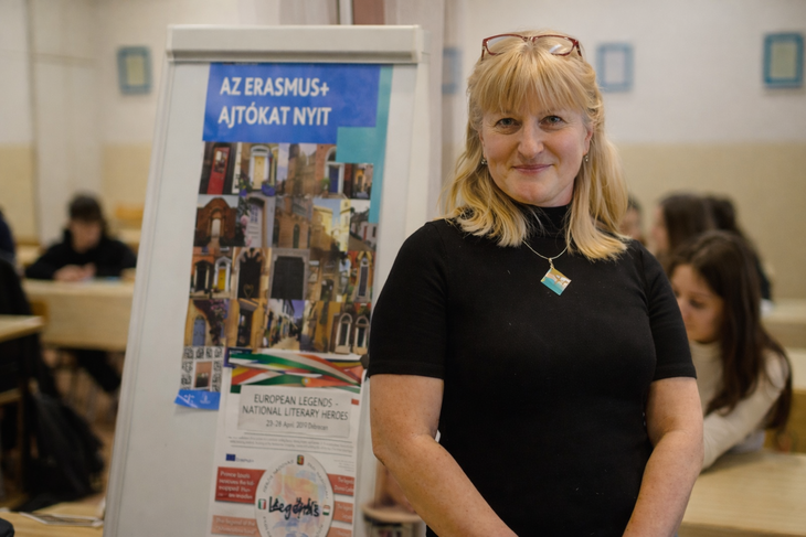 Szőke hölgy fekete felsőben tanteremben áll mögötte Erasmusplusz ajtókat nyit felirat