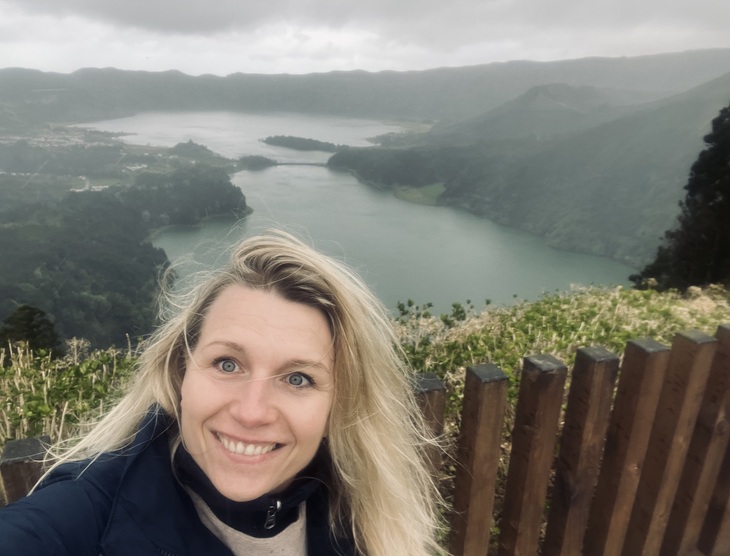 Szőke hölgy barna kerítés előtt fotózkodik egy folyóval a háttérben