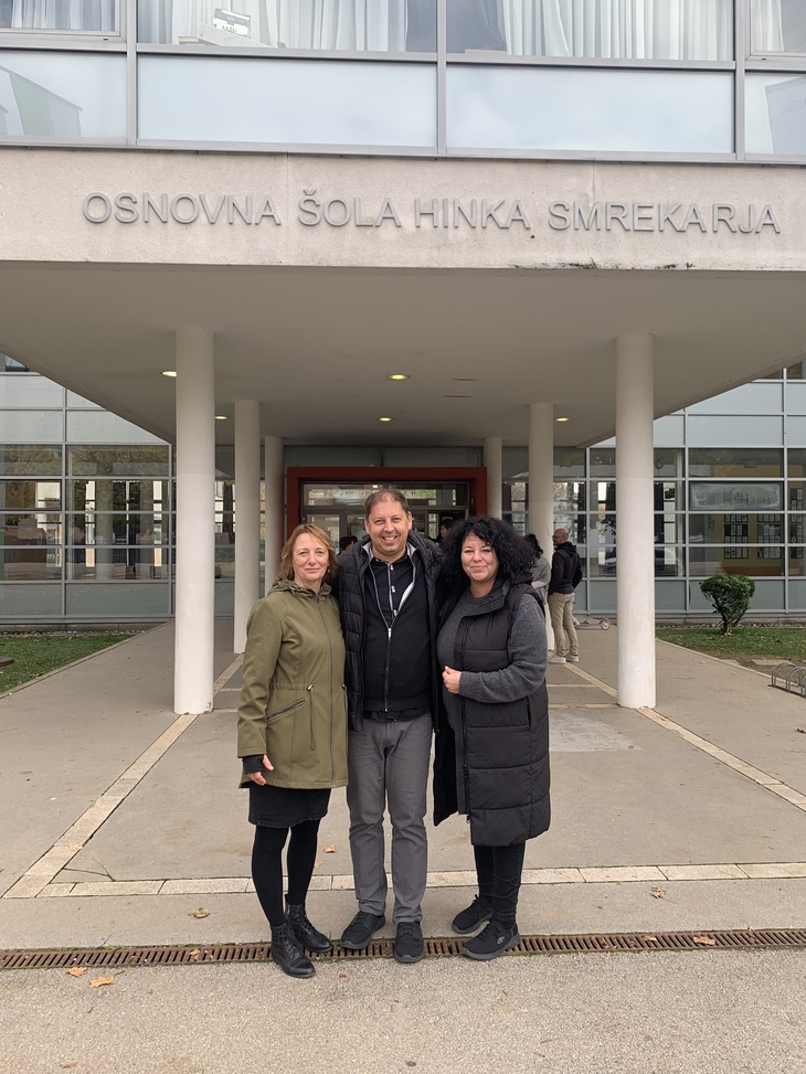Két nő és egy férfi kabátban állnak egy szlovén általános iskola bejáratánál
