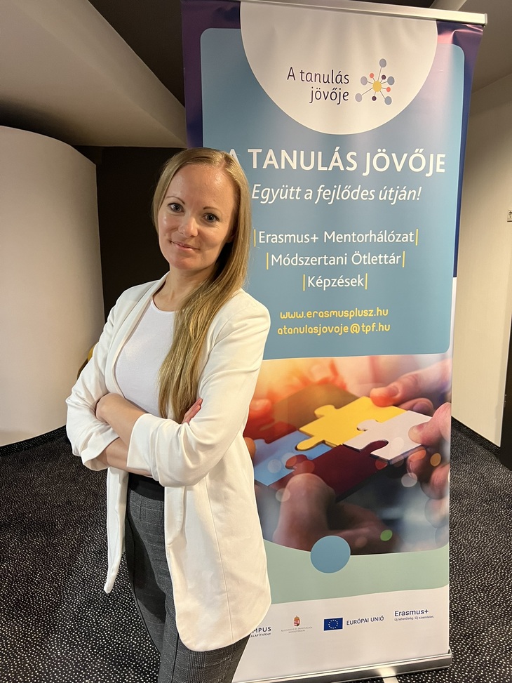 Fiatal hölgy fehér zakóban karba tett kézzel áll egy A tanulás jövője feliratú plakát előtt
