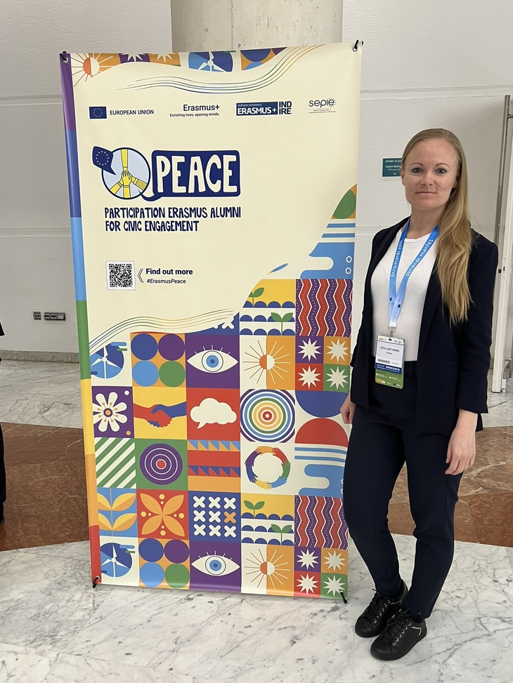 elegáns fiatal nő áll egy Erasmusplusz eseményt hirdető plakát előtt