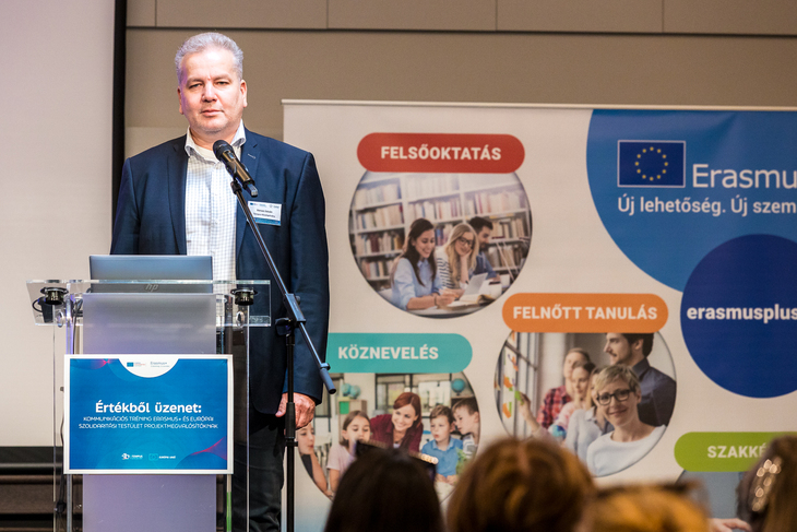 Öltönyös úr egy mikrofon előtt mond beszédet háttérben Erasmusplusz felirat és logó