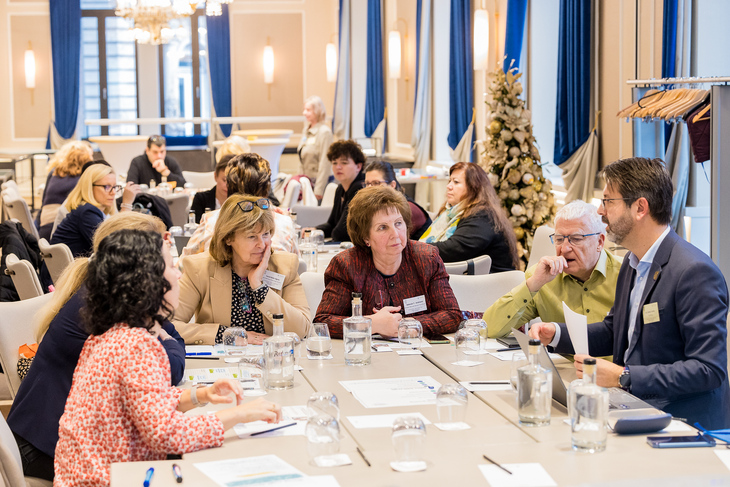 Elegánsan öltözött férfiak és nők asztalnál ülve beszélgetnek egy karácsonyi hangulatú teremben